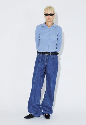 Persona che indossa un cardigan azzurro chiaro a coste con bottoni, jeans blu scuro larghi con cintura nera, scarpe nere appuntite e occhiali da sole rettangolari neri.