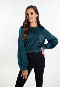 Teal satijnen blouse met lange, gepofte mouwen, verzamelde zoom en een zachte textuur. Gecombineerd met zwarte, aangesloten broeken en accenthardware.