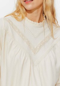 Haut en crème texturé avec un col ruffled, présentant des détails en dentelle en forme de V et un tissu froncé pour une coupe lâche.