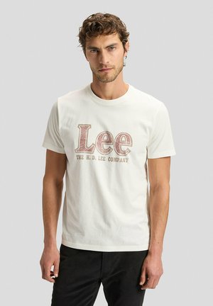 Mann mit kurzen, lockigen Haaren trägt ein weißes T-Shirt mit der Aufschrift "Lee THE H.D. LEE COMPANY" in großen Buchstaben und steht vor einem schlichten Hintergrund.
