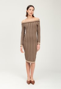 Eine Frau trägt ein figurbetontes, schulterfreies, langärmliges braunes, gemustertes Midi-Kleid und spitze braune Schuhe, während sie vor einem schlichten Hintergrund steht.