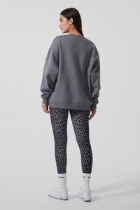 Femeie văzută din spate, purtând un hanorac gri larg, colanți negri și gri cu imprimeu leopard, șosete albe și adidași albi pe un fundal simplu.