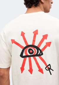 Άνδρας που φοράει λευκό πουκάμισο με μαύρο και κόκκινο γραφικό ενός ματιού περιβαλλόμενο από οκτώ κόκκινους βέλη που δείχνουν προς τα έξω στην πλάτη.