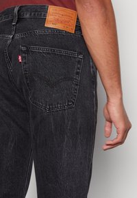 Jeans de mezclilla negra con corte recto, que presentan una etiqueta de cuero en la cintura y una sutil textura descolorida en la tela.