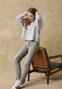 Grau gestrickter Pullover mit Rippstruktur, kombiniert mit hellgrauen, figurbetonten Jeans. Das Model trägt weiße Sneaker mit schwarzen Streifen. Im Hintergrund befindet sich ein Holzstuhl.