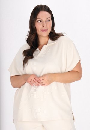 Lys beige top med et tekstureret geometrisk mønster, korte ærmer og en notch-krave. Sideslidser og en afslappet pasform.