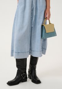 Nainen, joka on pukeutunut vaaleansinisiin leveisiin farkkuihin ja mustiin polvipitkiin saappaisiin, pitää kädessään kaksiväristä sinisen ja beigein käsilaukkua.