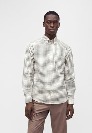 Homme portant une chemise blanche à boutons avec un motif en grille noire et un pantalon marron clair, debout devant un fond blanc uni.