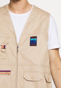 Beige väst med flera fickor och dragkedja fram. Har färgglada mönster på fickorna och en blå bergsillustration i ett patch.