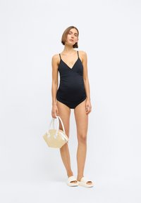 Femeie însărcinată stând într-un costum de baie de maternitate negru, ținând o geantă din paie și purtând sandale albe pe un fundal simplu.