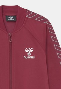 Bordeaux kleurige zip-up jasje met geribde kraag, voorzien van een wit geborduurd logo en gestructureerde grijze accenten langs de schouders in een Chevron patroon.