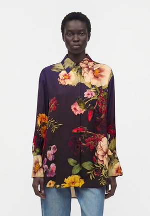 Femme portant une chemise boutonnée violet foncé avec de grandes impressions florales multicolores, associée à un jean bleu clair, debout devant un fond uni.