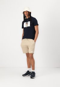 Camiseta navy con estampado gráfico, pantalones cortos beige con cordón, sombrero de cubo blanco y zapatillas deportivas negras. Outfit sencillo y casual.