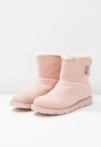 Paire de bottines en daim rose clair avec doublure blanche duveteuse et un bouton décoratif sur le côté, posées sur une surface blanche.