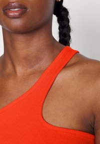 Červený žebrovaný top bez rukávů se širokým, asymetrickým ramínkem, s hladkým výstřihem a texturovanou látkou na neutrálním pozadí.