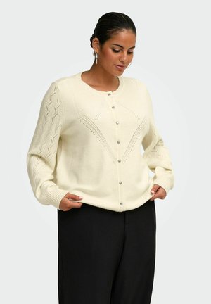 Vrouw draagt een crèmekleurige gebreide cardigan met knopen en met patroon op de mouwen en een zwarte broek, staand tegen een effen lichte achtergrond.
