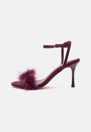 Sandale à talon haut violette avec bride de cheville et détail en plumes duveteuses sur la bride des orteils, présentée sur fond blanc.