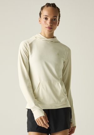 Young person with braided hair wears light beige hooded long-sleeve top and black shorts, standing against a plain background.