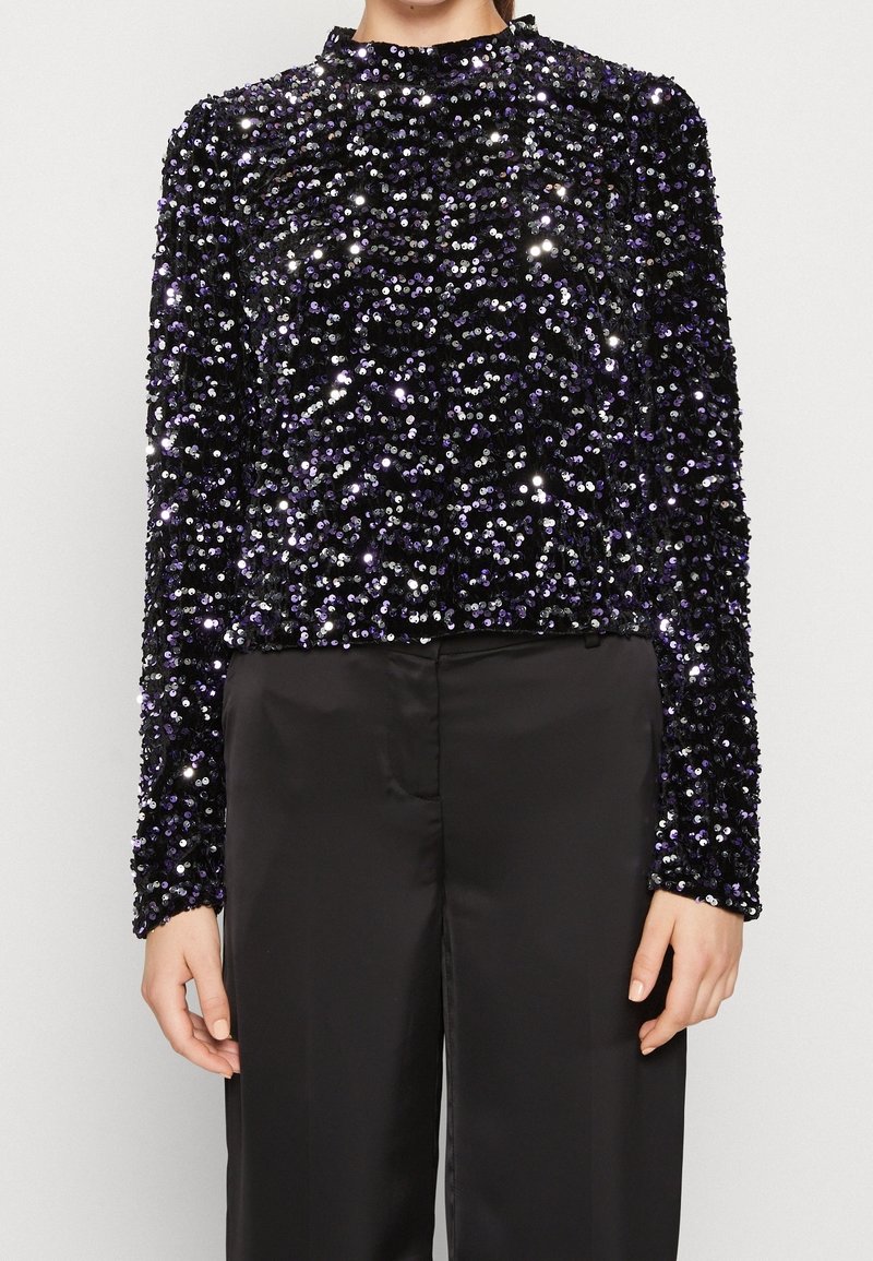 Personne porte un haut noir à manches longues couvert de sequins noirs et violets brillants, associé à un pantalon noir uni taille haute.