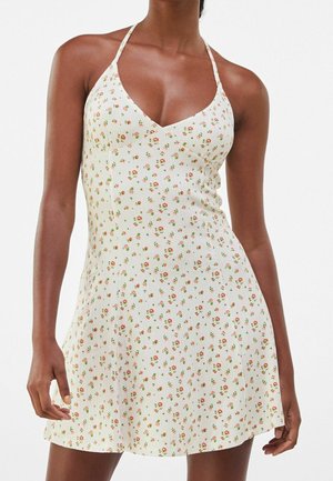 Nainen pukeutuneena valkoiseen halterneck-mekkoon, jossa on pieni oranssi- ja vihreä kukkakuvio, mekko istuu vyötäröltä ja levenee hieman helman kohdalla.