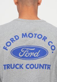Siva pamučna majica s plavim grafičkim printom na kojem je natpis "FORD MOTOR CO." i "TRUCK COUNTRY", okrugli izrez i kratki rukavi.