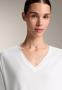Bílý tričko s V výstřihem vyrobené z hladkého, lehkého materiálu. Má uvolněný střih s krátkými rukávy a minimálním prošíváním.