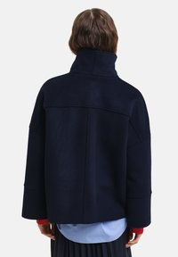 Ναυτικό oversized μπουφάν από μείγμα μάλλινου υφάσματος με ψηλό γιακά, χαμηλούς ώμους και ραφές, φορεμένο πάνω από ένα ελαφρύ γαλάζιο ριγέ πουκάμισο.