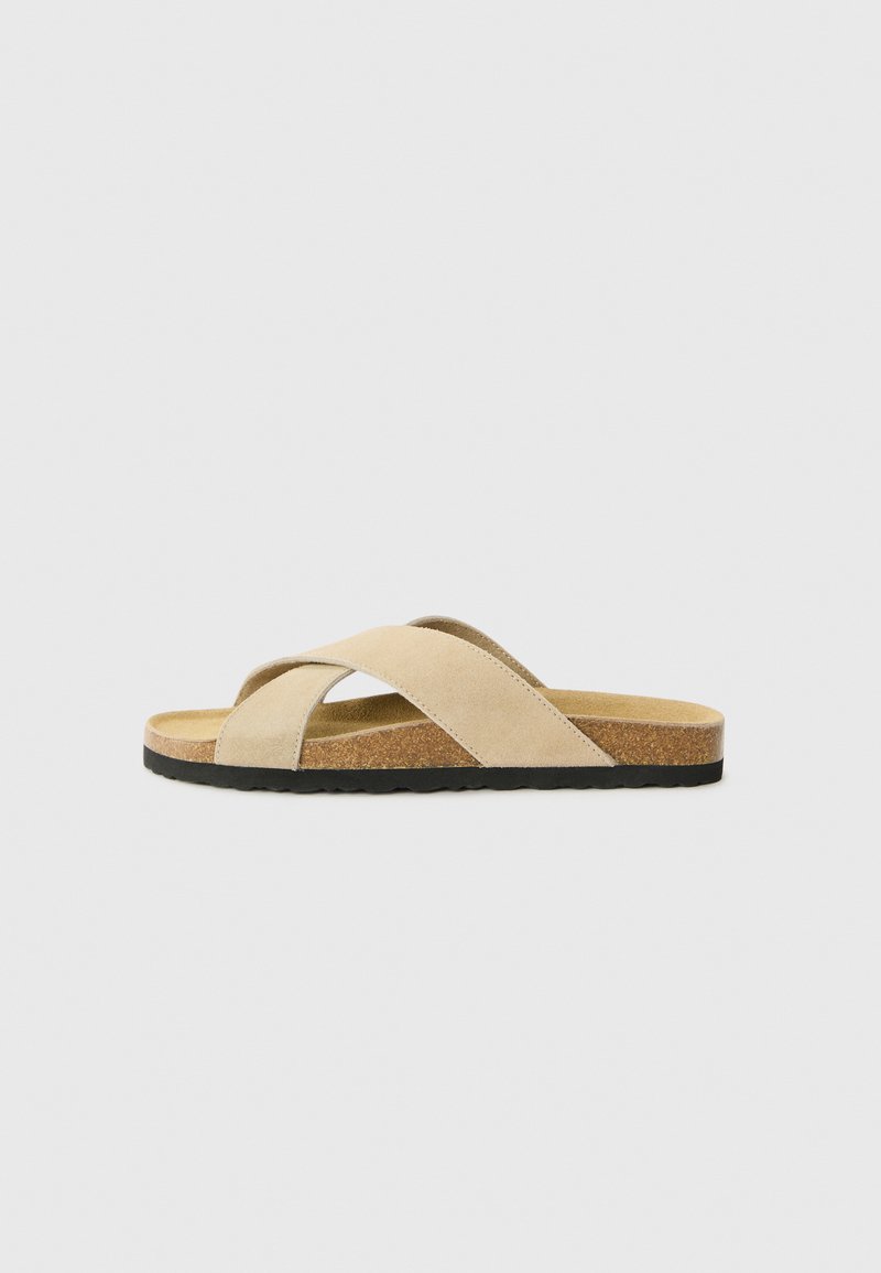 Sandale bej cu curea încrucișată, cu superior din suede, talpă din plută și talpă din cauciuc texturat negru, având un design plat și detalii minime.
