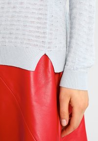 Suéter de punto azul claro con un patrón texturizado, que presenta un dobladillo acanalado y detalle de costura lateral, combinado con una falda de cuero rojo brillante.