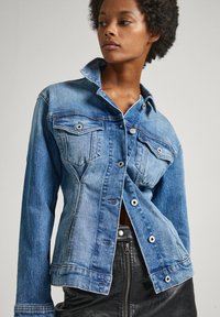 Denim jacket in medium wash blue, featuring two chest pockets, snap buttons, and a structured collar; worn open over a black leather skirt.