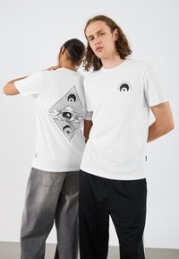 Hvite bomull T-skjorter med svarte grafiske design. Den ene har et øye-motiv foran; den andre viser et stort trekantdesign på baksiden.