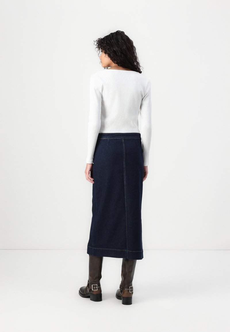 Weißes, geripptes Langarmoberteil, kombiniert mit einem dunkelblauen Jeans-Midirock, der mit Nähakzenten und einer geraden Silhouette versehen ist. Braune knielange Stiefel.