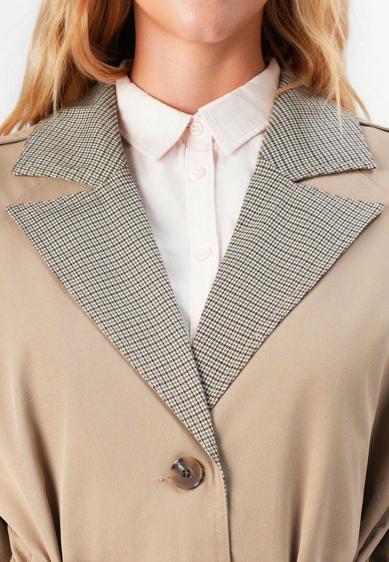 Gros plan d'une personne portant un manteau beige avec des revers à carreaux et une chemise blanche à boutons en dessous.