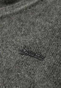 Tissu gris texturé présentant un motif tissé subtil et un logo "Superdry" brodé dans une teinte plus foncée.