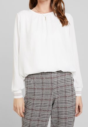 Kobieta ubrana w białą bluzkę z długim rękawem i marszczonymi mankietami, zestawioną z wysokim stanem czarno-biało-czerwonych spodni w kratę.