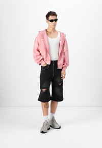 Lys pink oversized hoodie, hvid tanktop, sorte slidte shorts med frynsede kanter, grå sportssko og mørke solbriller.