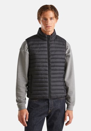 Giovane uomo con indosso un gilet trapuntato nero senza maniche sopra una felpa grigia e jeans scuri, in piedi davanti a uno sfondo chiaro e uniforme.