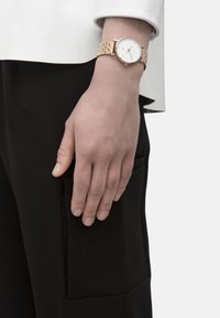 Gouden polshorloge met een ronde witte wijzerplaat, schakeltjesarmband en eenvoudige uurmarkeringen, gedragen op een neutraal getinte hand tegen een zwarte stof.