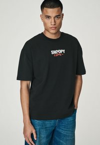 Sort bomulds t-shirt med "SNOOPY" grafik i hvid og rød. Korte ærmer, afslappet pasform, rund hals, parret med blå denim jeans.