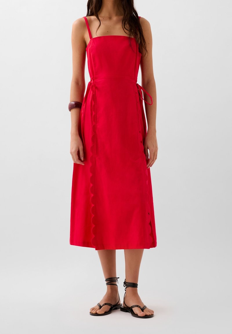 Robe midi rouge à fines bretelles, avec un détail noué sur le côté, un ourlet festonné et un tissu léger. Portée avec des sandales plates noires.
