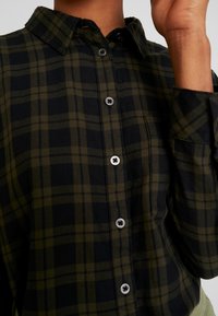 Chemise à carreaux verts et noirs avec un col boutonné, des boutons noirs et une poche poitrine. Le tissu semble doux avec une finition texturée.