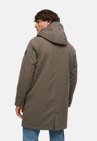 Zaļš, ar kapuci parka jaka ar gludu tekstūru, nedaudz garāka piegriezuma, sānu kabatām un melnu uzlīmi kreisajā plecā.