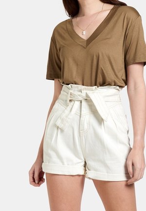 Kobieta ubrana w brązową krótkawą bluzkę z dekoltem w szpic, włożoną w białe, wysokim stanem szorty typu paperbag z wiązanym paskiem.