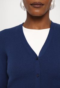 Mörkblå ribbad kofta med V-hals, med knappstängning fram över en vit skjorta, som framhäver textur och färgkontrast.