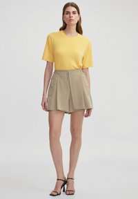 Camiseta amarilla de manga corta combinada con pantalones cortos beiges que tienen bolsillos laterales y cordón. La modelo lleva sandalias de tacón negro.