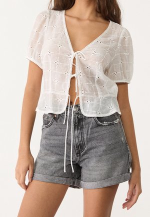 Woman wearing a white short-sleeve eyelet blouse with front ties and high-waisted rolled-hem gray denim shorts.