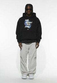 Sudadera negra con el texto "juventud desperdiciada en los jóvenes" y un gráfico de la Tierra azul, combinada con pantalones baggy de color gris claro y zapatillas blancas.