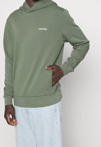 Zelená bavlněná mikina s kapucí, má přední klokankovou kapsu a jemné bílé logo Calvin Klein na hrudi. Hladká textura a uvolněný střih.