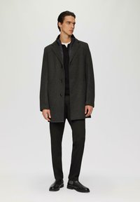 Grau melierter Wollmischmantel mit Reverskragen, drei Knöpfen und wattierter Innenauskleidung, kombiniert mit schwarzen trousers und Lederschuhen.
