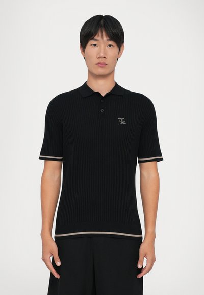 Hombre joven de pie, mirando hacia adelante, vestido con una camiseta polo negra de manga corta con ribetes beige en las mangas y el dobladillo.